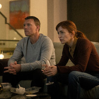 Two people sit on a couch in a dimly lit living room, looking intently at something offscreen. They have takeout containers and drinks on the table, suggesting a casual night at home. Warm lighting creates a cozy and focused atmosphere.