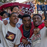 Three young men pose for a selfie at an outdoor event with a group, some wearing college-themed shirts, festive decorations and people with pom-poms in the background, creating a lively and celebratory atmosphere.