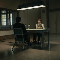 Two people sit across from each other at a table in a dimly lit interrogation room, with a large mirror on the wall reflecting the scene and a microphone on the table, creating a serious and tense atmosphere.