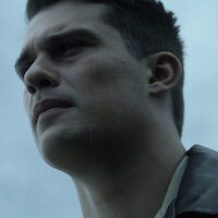 Close-up of a young man with short dark hair looking resolute, outdoors under overcast sky, wearing a jacket.