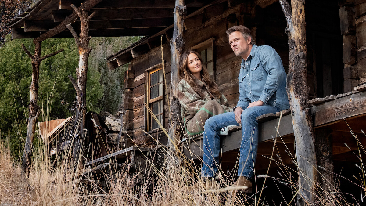 Man and woman sitting on porch of rustic wooden cabin in nature, surrounded by tall dry grass and trees, sharing a quiet moment outdoors.
