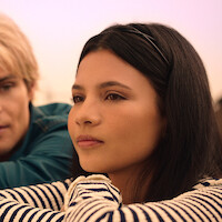Young woman with long dark hair and striped shirt outdoors, resting her chin on her arms, with a man in a blue shirt beside her, both gazing thoughtfully into the distance in soft natural light.