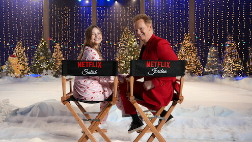 Two people smiling while sitting on director’s chairs with Netflix logos in a festive winter set decorated with snow, Christmas trees, twinkling lights, and holiday decorations, creating a cheerful holiday atmosphere.