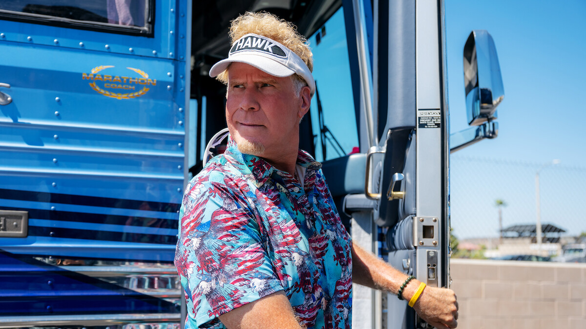Man in a tropical shirt and visor stands at the doorway of a blue luxury bus on a sunny day, holding the door handle, with a fence and clear sky in the background.