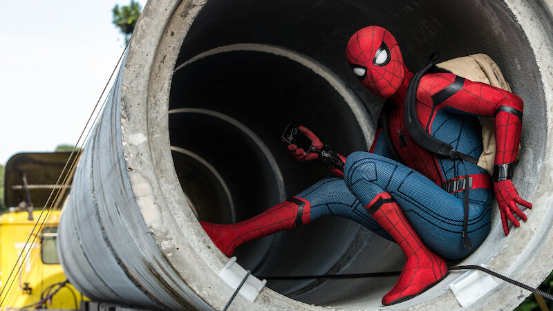 A person in a Spider-Man costume crouches inside a large concrete pipe outdoors, holding a device and wearing a backpack, with construction vehicles and trees visible in the background.