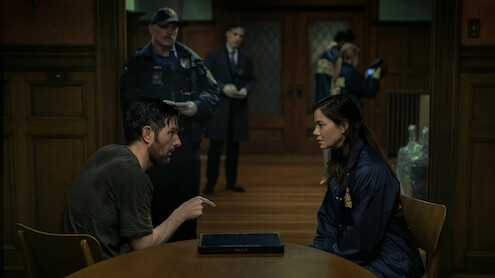 A man and a woman sit across from each other at a table in a dimly lit police interrogation room while officers and investigators work in the background.