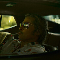 Man wearing sunglasses sits in the driver’s seat of a car, viewed from the side window, with sunlight filtering in. The car interior has a retro style and relaxed vibe.