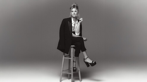 Meghann Fahy sits on a stool in a chic black suit with white button up blouse with legs crossed and platform heels.
