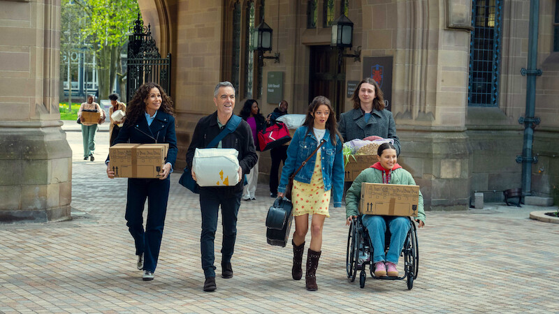 Five people carrying boxes and bags, including one in a wheelchair, walk together outside a historic stone building with arches and lanterns, suggesting a college or university move-in day.