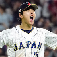 Shohei Ohtani celebrating on the field, arms raised and mouth open in excitement, wearing a white pinstripe jersey and glove, with a blurred crowd in the stadium background.