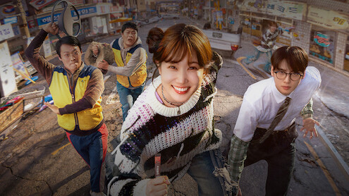 Five people, including a smiling woman in a colorful sweater and a man in glasses, run on a damaged street in a city neighborhood with cracked pavement and rundown storefronts, suggesting urgency or disaster in an urban setting.