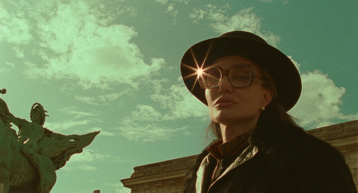 Angelina Jolie as Maria Callas stands in front of a cloudy sky, wearing a black hat and large sunglasses that reflect the sunlight back to the camera.