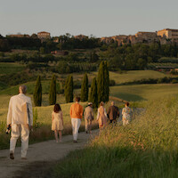 A group of people walk down a country path through green fields at sunset, heading toward a distant hilltop village, evoking a peaceful and warm atmosphere.