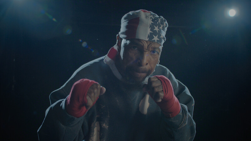 A male boxer wearing red hand wraps and a USA flag bandana poses in a fighting stance under dramatic lighting in a dark gym or training environment.