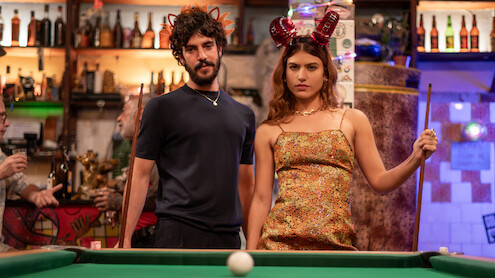 Um homem de camiseta preta, barba e cabelos grandes, e uma mulher de cabelos longos e castanhos, vestindo um vestido amarelo, se posicionam na frente de uma mesa de sinuca com a bola branca centralizada no primeiro plano