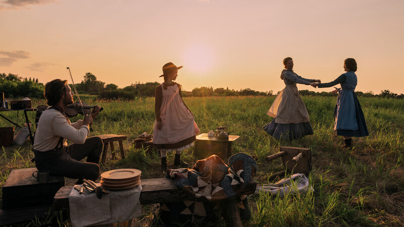 Pessoas usando roupas do século 19 reunidas ao pôr do sol. Um homem toca violino enquanto três mulheres dançam em um gramado com mesas rústicas e bancos de madeira, criando um clima nostálgico e rural.