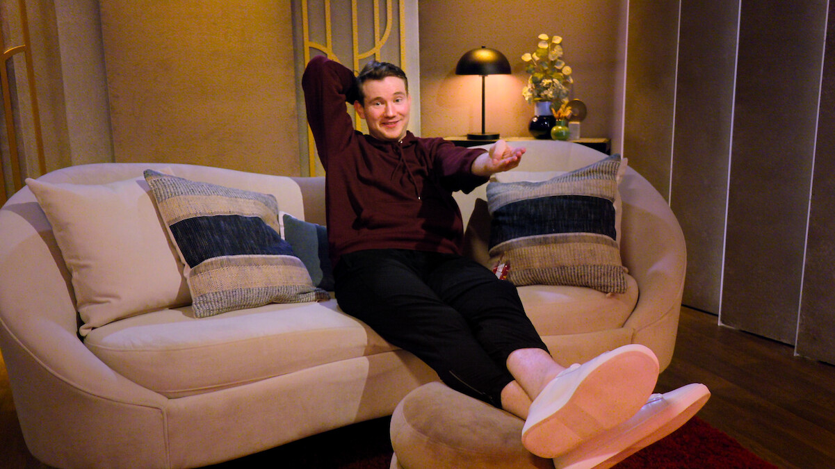 Person relaxing on a beige couch with blue and gray pillows in a cozy, warmly lit living room with a lamp, flowers, and decorative items on a side table; modern, comfortable setting.