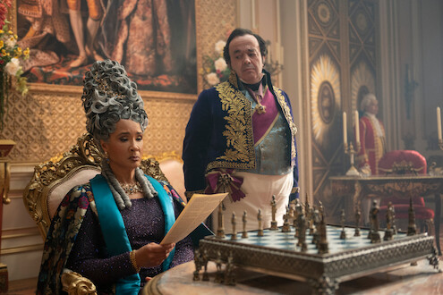 A regal woman in elaborate period attire reads a letter while seated at an ornate chess table in a lavish, opulent room; a man in formal clothing stands behind her, with rich decorations and candelabras in the background.