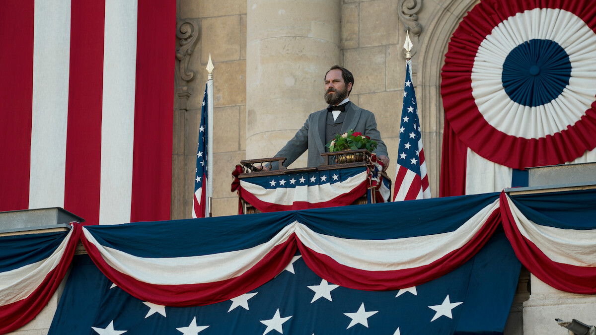 A man in historical attire stands on a decorated balcony with American flags and patriotic bunting, delivering a speech in a formal, historic setting. The mood is solemn and ceremonial.