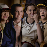 Gaten Matarazzo and Millie Bobby Brown from Stranger Things, shown as both kids and adults, seated in interview settings, reacting and smiling.
