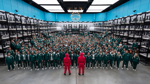 Hundreds of people in green tracksuits stand in rows inside a large, brightly lit room with bunk beds, facing two guards in pink uniforms. 