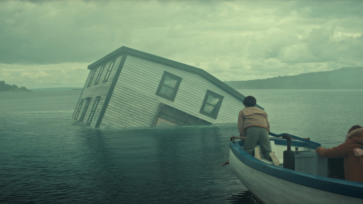 Two people in a small boat approach a partially submerged house tilted in the middle of a misty lake under a cloudy sky, with forested hills in the background.