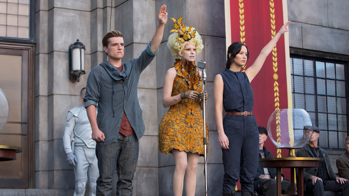 Three people stand in front of a stone building, two raising one hand; one wears a grey shirt, another a sleeveless outfit, and the third is in an elaborate orange dress at a microphone. Others are seated nearby, and a large banner hangs.
