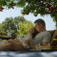 A couple relaxes together on outdoor lounge chairs under a shaded arbor with flowers, surrounded by green trees and a garden setting. A pitcher of water and glasses are on a nearby table, suggesting a peaceful afternoon.