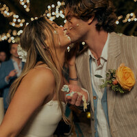 Bride and groom with cake on their faces share a kiss at an outdoor wedding reception. String lights create a festive atmosphere, while guests watch in the background. The groom wears a flower boutonniere.