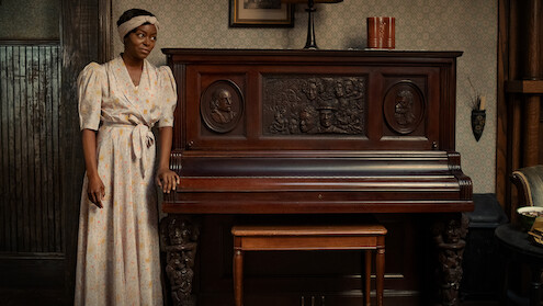Berniece Charles (Danielle Deadwyler) stands in a white dress at an intricately carved wooden piano. 