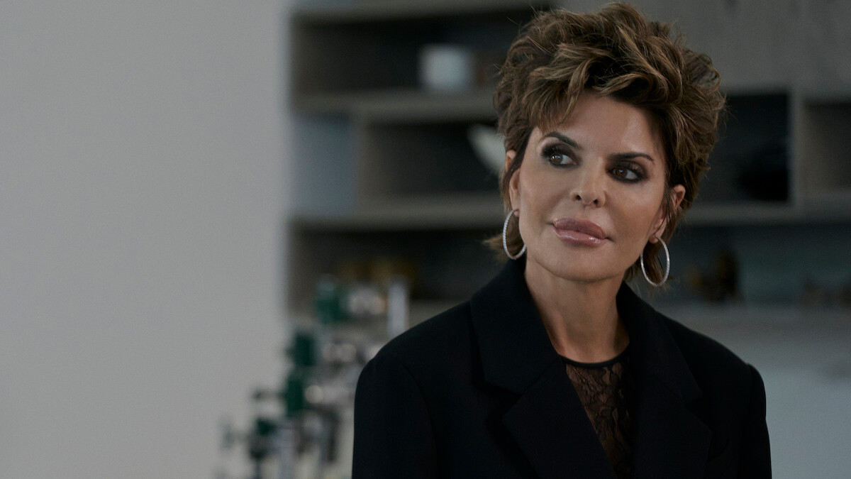 Woman with short styled hair and hoop earrings wearing a black blazer in a modern indoor setting with shelves and bottles blurred in the background, looking thoughtfully to the side.