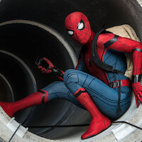 A person in a red and blue superhero costume crouches inside a large concrete pipe, holding a device and wearing a beige backpack in an urban, industrial setting.