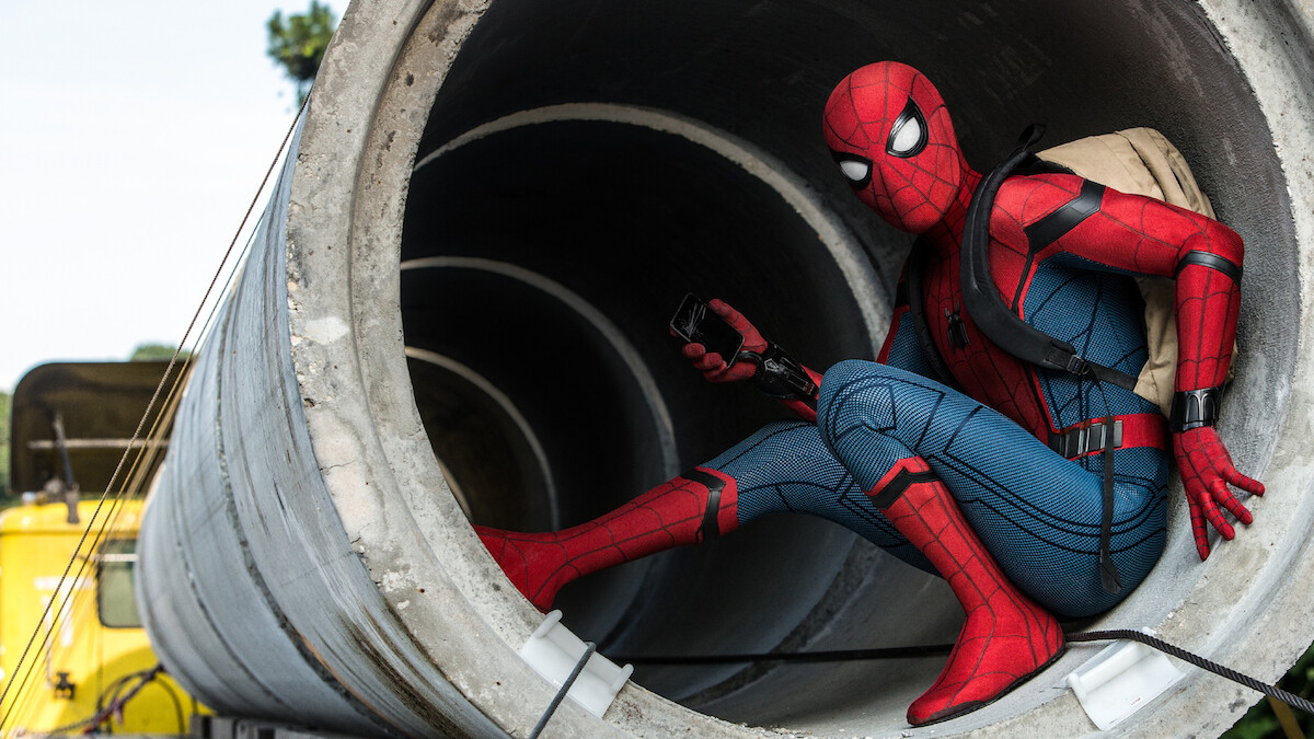 A person in a Spider-Man costume crouches inside a large concrete pipe outdoors, holding a device and wearing a backpack, with construction vehicles and trees visible in the background.