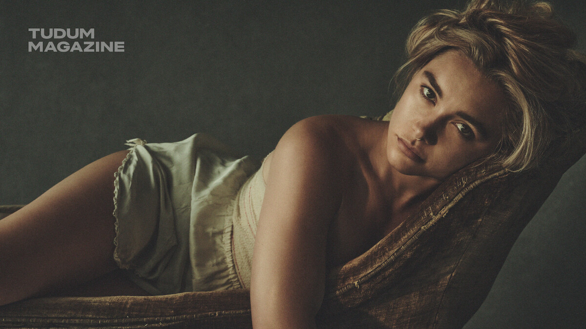 Woman with blonde hair in light clothing reclines on a vintage chair, looking directly at the camera in a dimly lit, moody setting. Tudum Magazine logo appears in the upper left corner.