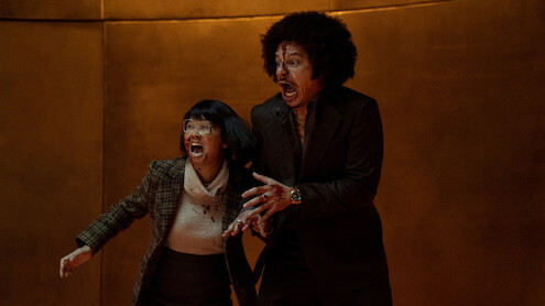 Two people with shocked expressions, one wearing glasses and plaid, the other in a dark suit with an afro, stand together in a dimly lit room with gold-colored walls, appearing frightened or alarmed by something off-camera.