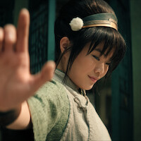 Miyako as Toph, with long hair in a bun, wearing a headband and traditional attire, smiling gently, extends hand outward confidently.