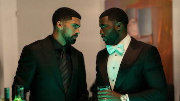 Two men in formal black suits stand close, engaged in a serious conversation at an indoor event. One holds a drink, bottles are visible on the table, and ambient green lighting enhances the elegant, upscale setting.