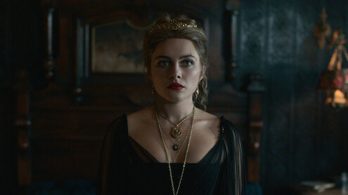 Uma mulher usando um vestido preto e diversos colares em uma sala com pouca iluminação que tem móveis de madeira escura e decoração antiga. Ela olha para frente com uma expressão séria.