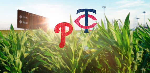 Baseball field scoreboard and stadium lights in the background, lush green cornfield in foreground, with Philadelphia Phillies and Minnesota Twins logos superimposed in the center, sunlight shining through.
