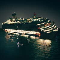 A large cruise ship listing heavily at night, brightly lit, with lifeboats and rescue craft on the water nearby, indicating an emergency situation at sea.