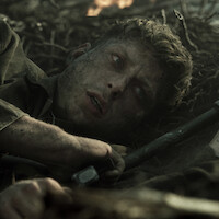 A weary soldier with a dirty face lies on the ground in a battlefield setting, surrounded by debris and dirt, looking upward with a tense, anxious expression. The environment is dark and smoky, hinting at an ongoing conflict.