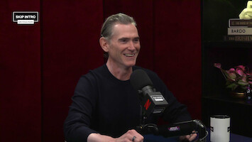 A smiling man sits in a podcast studio with a microphone, dark red curtains, and shelves with books and flowers behind him. The mood is relaxed and conversational, suggesting a friendly interview setting.
