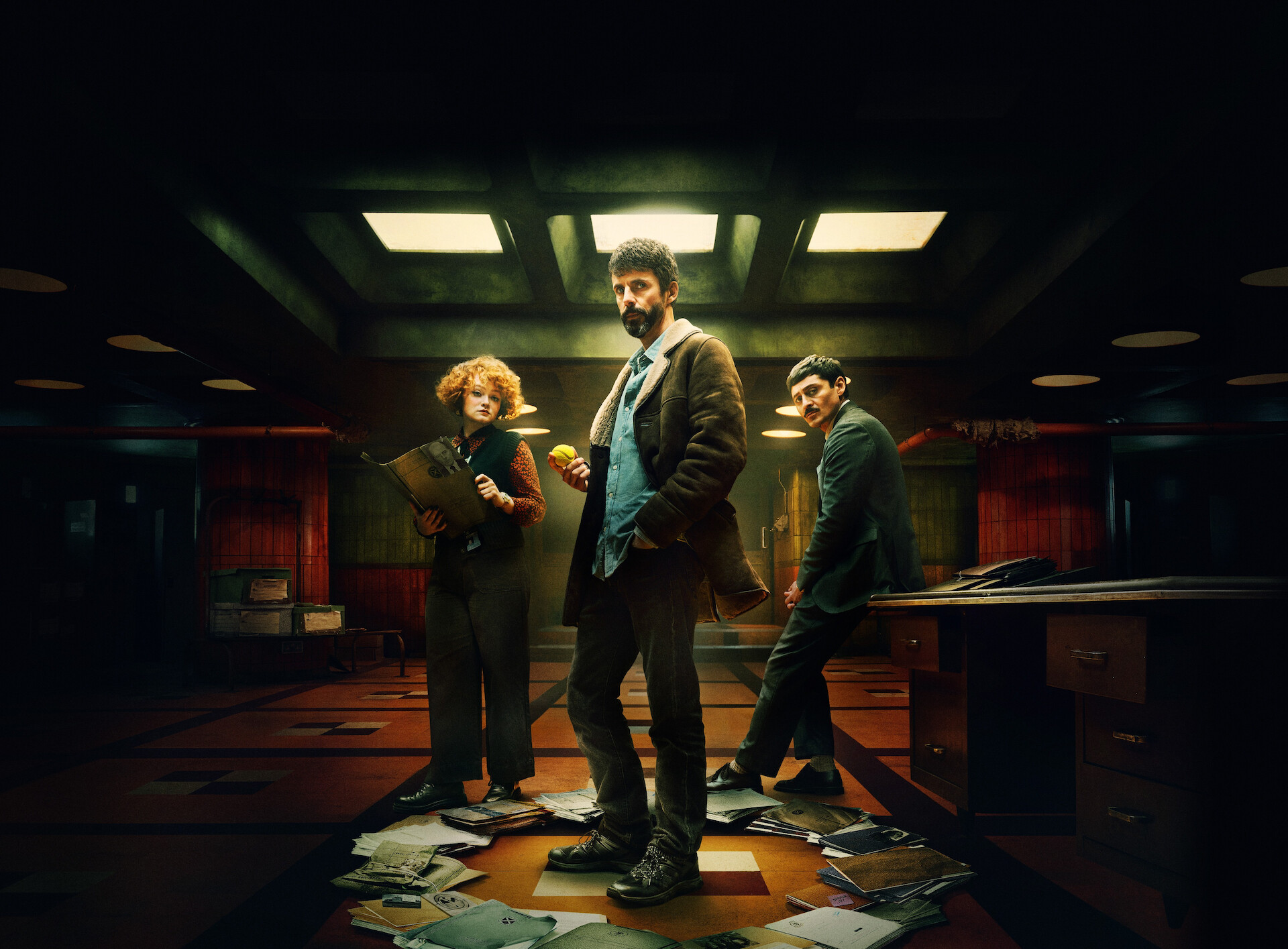 Dept. Q - The cast of Dept Q standing in an office with paperwork scattered around them in a circle. 