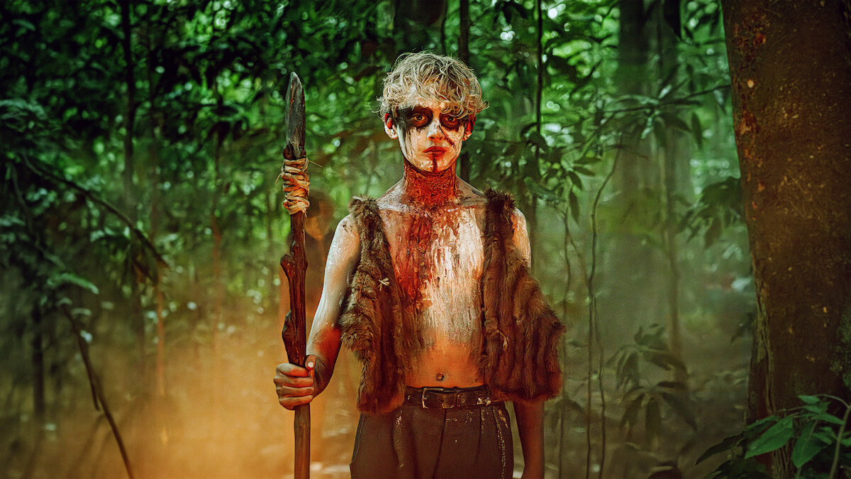 A person with painted face and fur vest stands holding a spear in a dense, misty, green forest, suggesting a primitive or survival setting.
