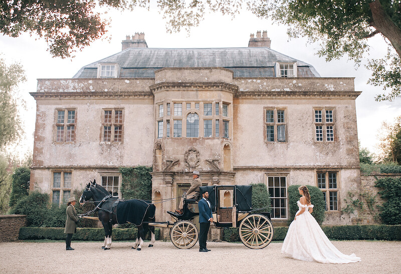 Bride in a white gown stands near a vintage horse-drawn carriage outside an elegant, historic mansion. Lush greenery frames the scene, evoking a romantic, fairytale atmosphere. The mood is dreamy and regal.