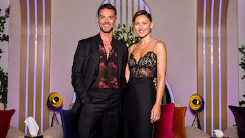A man in a black suit and a woman in a black lace dress stand together smiling in a modern lounge with colorful cushions, round lights, plants, and decorative vertical lines in the background.