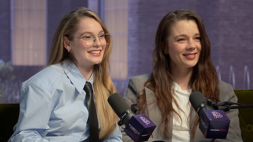 Two women in business attire smile and sit on a couch during an indoor podcast or interview, each speaking into a microphone with a blurred cityscape visible through large windows in the background.