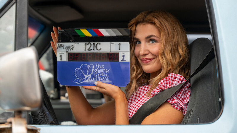 Smiling woman in a car holding a film clapperboard labeled "this summer will be different" wearing a red-checkered shirt and seatbelt, filming scene in bright daylight.