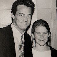 Man in a suit and tie smiling next to a young girl with long hair in a casual top, both posing indoors against a paneled wall in a black and white photograph.