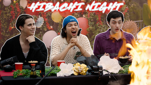 From left to right, Tanner Buchanan, Xolo Maridueña, and Gianni DeCenzo are seated at a dinner table, with flames rising dramatically on the right side.